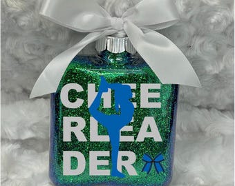 May include: A square glass ornament filled with green and blue glitter, topped with a white satin ribbon bow. The front features white text reading CHEERLEADER, with a blue silhouette of a cheerleader and a small blue bow graphic.