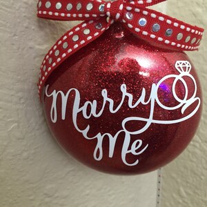May include: Red glitter ornament with the words "Marry Me" in white script. The ornament has a diamond ring graphic and is adorned with a red ribbon with silver dots. A festive proposal decoration.