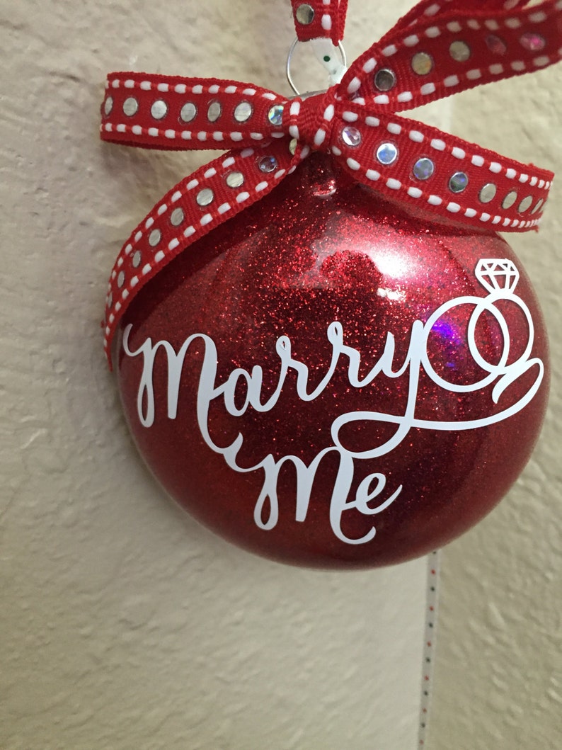 May include: Red glitter ornament with the words "Marry Me" in white script. The ornament has a diamond ring graphic and is adorned with a red ribbon with silver dots. A festive proposal decoration.