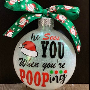 May include: Glittery Christmas ornament with the text "he Sees YOU When you're POOPING" and a Santa hat graphic. The ornament has a green ribbon with Santa Claus designs. The text "GLOW IN THE DARK" is at the bottom.