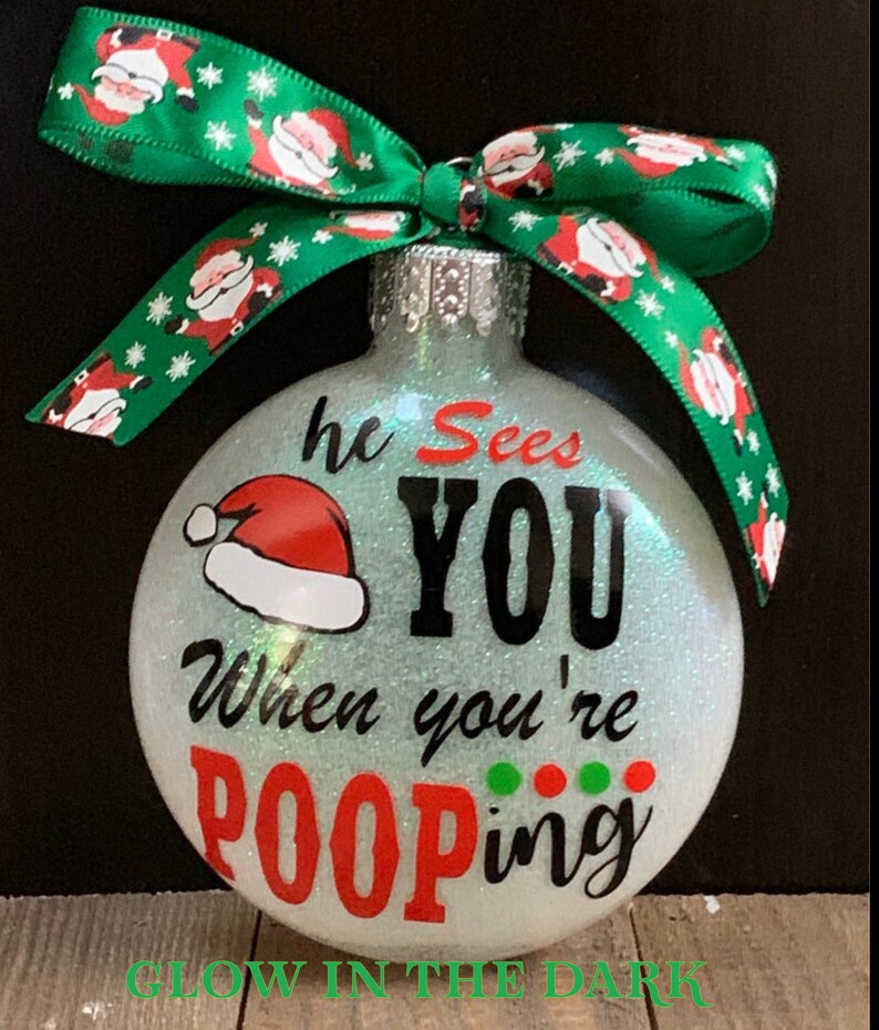 May include: Glittery Christmas ornament with the text "he Sees YOU When you're POOPING" and a Santa hat graphic. The ornament has a green ribbon with Santa Claus designs. The text "GLOW IN THE DARK" is at the bottom.