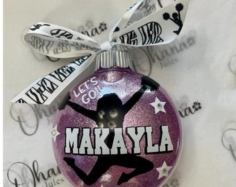 May include: A round purple glitter ornament featuring a black silhouette of a cheerleader jumping with pom-poms. The text reads LET'S GO! and MAKAYLA in white and black lettering. Three small white stars with rhinestones accent the design.