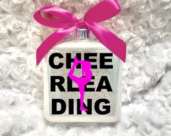 May include: A square, white glitter ornament featuring the black text CHEERLEADING with a bright pink silhouette of a cheerleader in a scorpion pose replacing the letter R, topped with a vibrant pink satin ribbon bow.