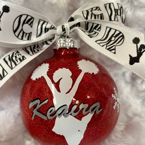 May include: A round red glitter Christmas ornament featuring a white silhouette of a cheerleader holding pom-poms, the name Keaira in silver script, and a small white snowflake. A white ribbon with black zebra print and the word CHEER is tied on top.