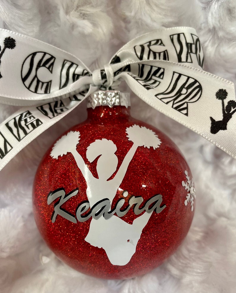 May include: A round red glitter Christmas ornament featuring a white silhouette of a cheerleader holding pom-poms, the name Keaira in silver script, and a small white snowflake. A white ribbon with black zebra print and the word CHEER is tied on top.
