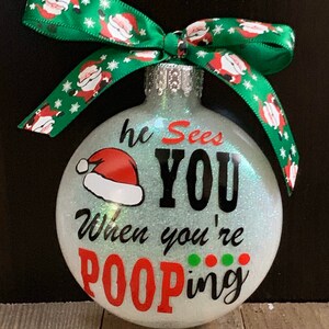 May include: Glittery Christmas ornament with the text "he Sees YOU When you're POOPing" in black and red. A Santa hat graphic is above the text. The ornament has a green ribbon with Santa Claus designs.