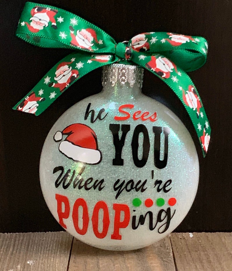 May include: Glittery Christmas ornament with the text "he Sees YOU When you're POOPing" in black and red. A Santa hat graphic is above the text. The ornament has a green ribbon with Santa Claus designs.
