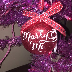 May include: A red glitter Christmas ornament with the words "Marry Me" in white script. The ornament is adorned with a red ribbon with white polka dots and silver embellishments. A diamond ring hangs below.