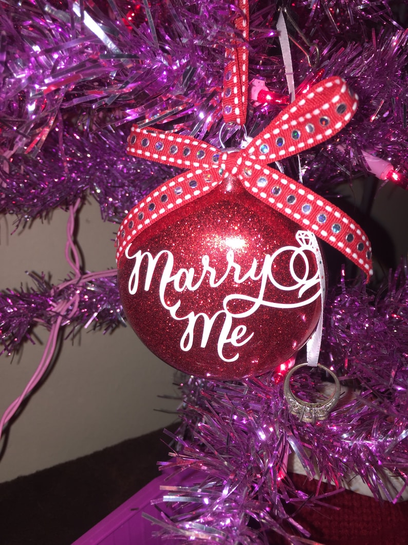 May include: A red glitter Christmas ornament with the words "Marry Me" in white script. The ornament is adorned with a red ribbon with white polka dots and silver embellishments. A diamond ring hangs below.