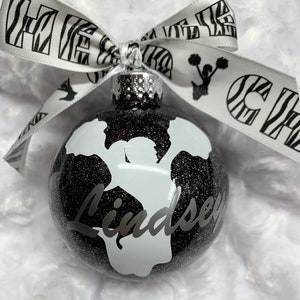 May include: A spherical black glitter ornament featuring a white silhouette of a cheerleader holding pom-poms and the name Lindsey in script. A white ribbon with black zebra print, cheerleading silhouettes, and the text CHEER is tied at the top.