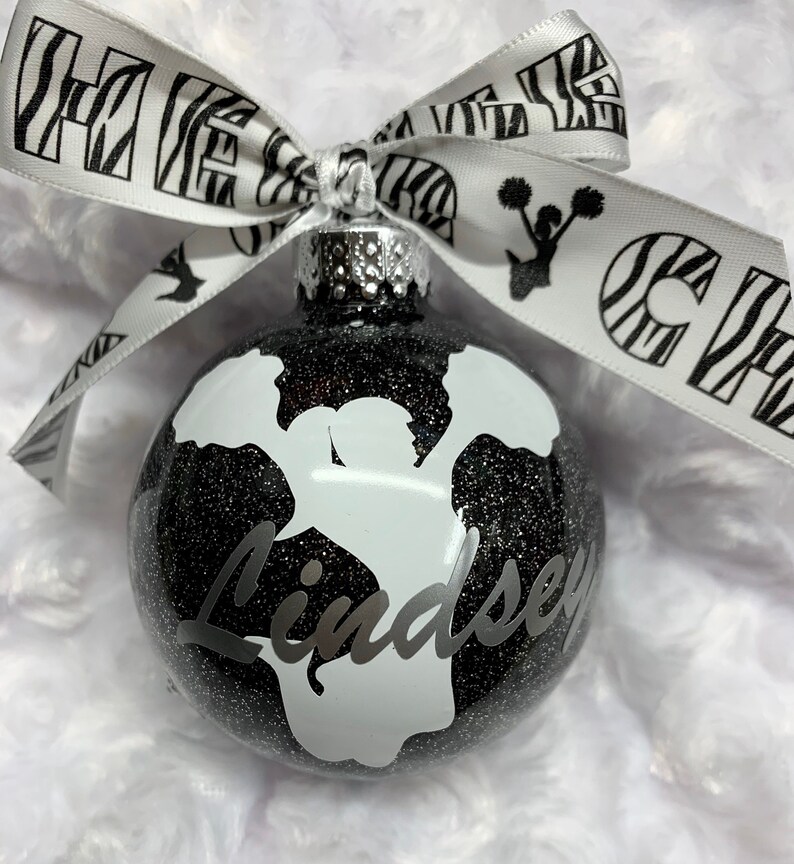 May include: A spherical black glitter ornament featuring a white silhouette of a cheerleader holding pom-poms and the name Lindsey in script. A white ribbon with black zebra print, cheerleading silhouettes, and the text CHEER is tied at the top.