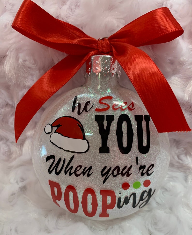 May include: Glittery Christmas ornament with a red ribbon bow. The ornament has the text "he Sees YOU When you're POOPING" with a Santa hat graphic. The text is in black and red.