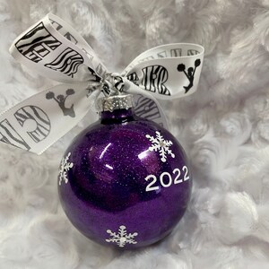 May include: A round purple glitter Christmas ornament featuring white snowflake decals and the year 2022 printed in white. A white ribbon with black zebra print and black cheerleader silhouettes is tied to the silver metal cap at the top.