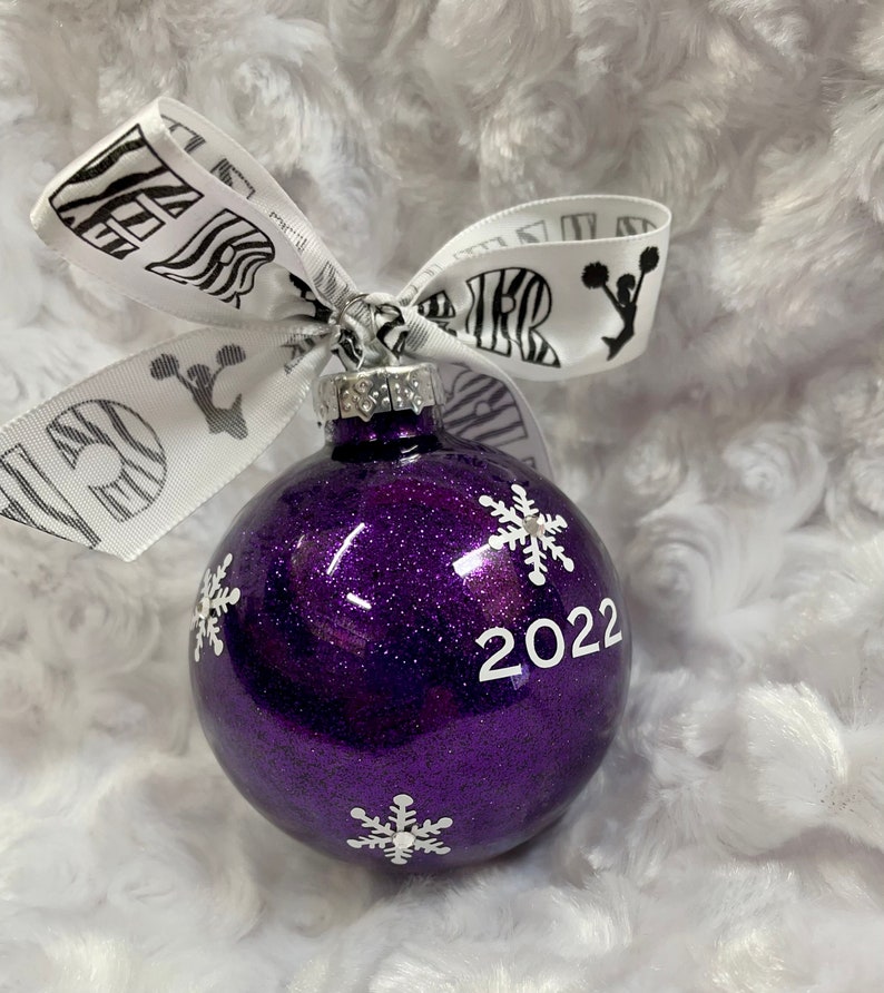 May include: A round purple glitter Christmas ornament featuring white snowflake decals and the year 2022 printed in white. A white ribbon with black zebra print and black cheerleader silhouettes is tied to the silver metal cap at the top.