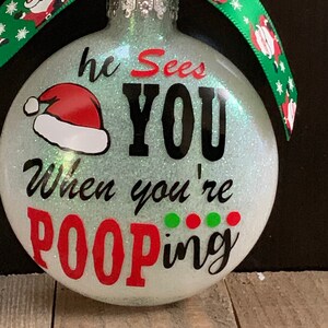 May include: Glittery Christmas ornament with the text "he Sees YOU When you're POOPING". The ornament has a Santa hat graphic and is decorated with a green ribbon with Santa Claus designs.