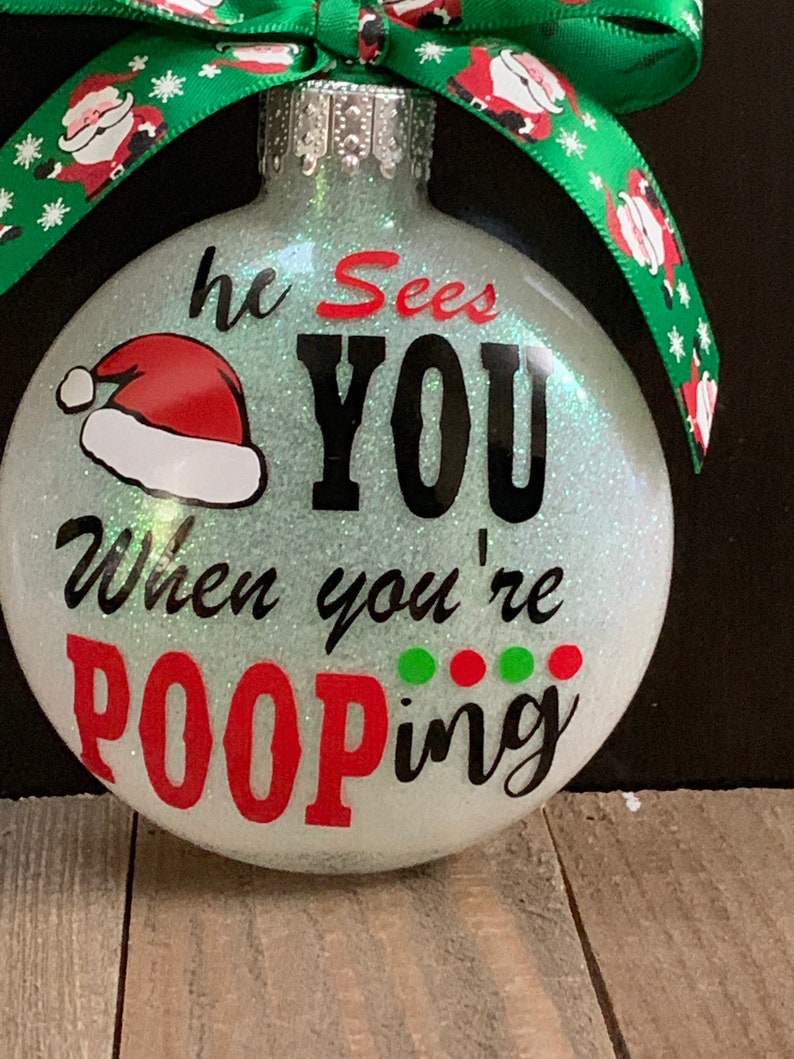 May include: Glittery Christmas ornament with the text "he Sees YOU When you're POOPING". The ornament has a Santa hat graphic and is decorated with a green ribbon with Santa Claus designs.