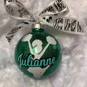 May include: A spherical green glitter Christmas ornament featuring a white silhouette of a cheerleader holding pom-poms. The name Julianne is printed in teal script across the center. A white ribbon with black zebra print and the word CHEER is tied to the top.