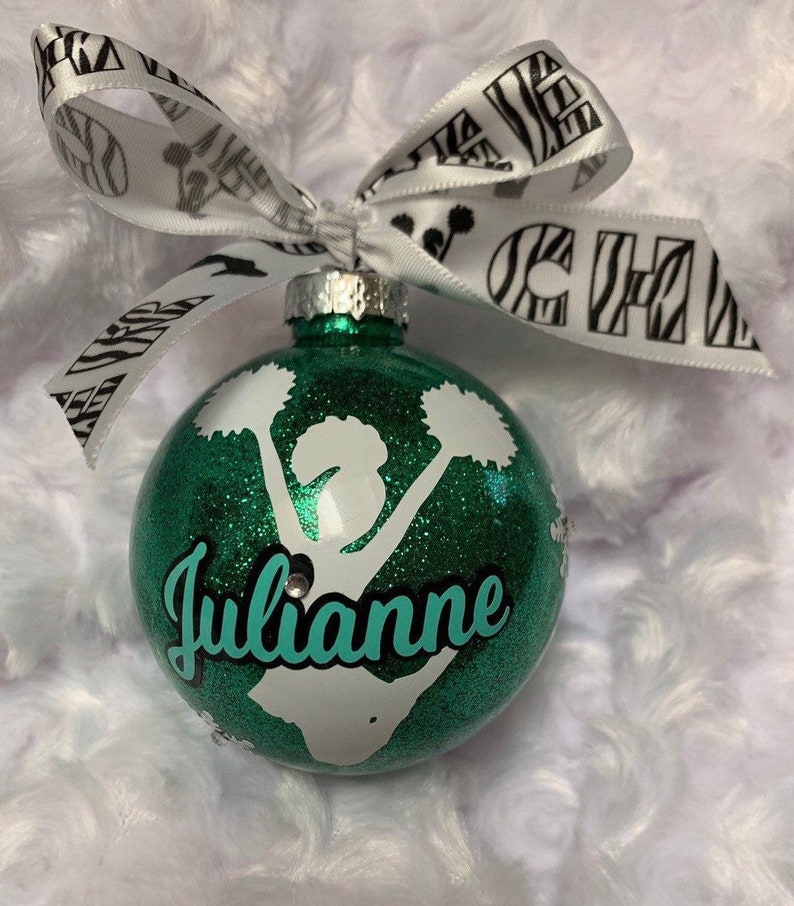 May include: A spherical green glitter Christmas ornament featuring a white silhouette of a cheerleader holding pom-poms. The name Julianne is printed in teal script across the center. A white ribbon with black zebra print and the word CHEER is tied to the top.