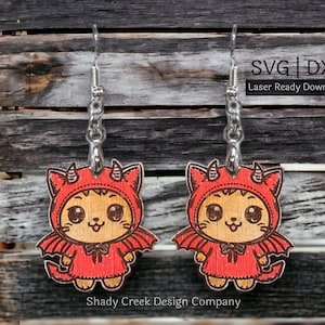 May include: A pair of laser-cut wooden earrings featuring a cute cartoon cat dressed as a devil. The cat is wearing a red hooded costume with horns and wings. The earrings are hanging from silver hooks.