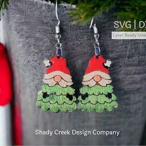 Może przedstawiać: Para drewnianych kolczyków w kształcie choinki z twarzą gnoma na każdym drzewie. Kolczyki są zielone, czerwone i białe. W obrazie widoczny jest tekst "SVG | DXF Laser Ready Download".