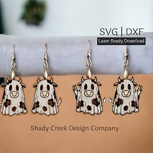May include: Four wooden cow ghost earrings with black and white spots. The earrings are hanging from silver hooks. The text "SVG | DXF Laser Ready Download" is at the top of the image. The text "Shady Creek Design Company" is at the bottom of the image.
