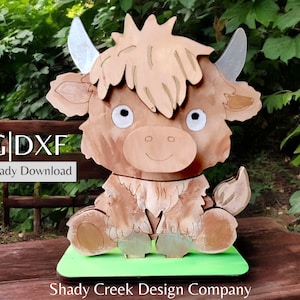 Puede incluir: Un recorte de madera cortado con láser de una vaca Highland marrón y blanca con una base verde. El recorte está etiquetado como "SVG|DXF Descarga lista para láser" y "Shady Creek Design Company".