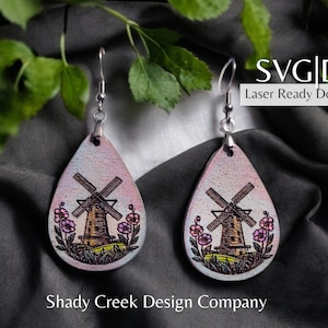 May include: A pair of dangle earrings with a pink and white teardrop-shaped design. Each earring features a small windmill with a brown roof and a green base, surrounded by pink flowers. The earrings are made of leather and have a silver hook.
