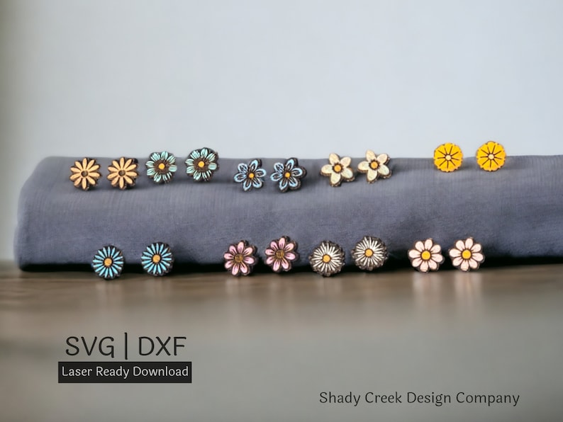 May include: A set of 12 laser cut wooden flower earrings in various colors, including yellow, blue, pink, and white. The earrings are arranged on a gray fabric surface. The text "SVG | DXF Laser Ready Download" and "Shady Creek Design Company" are visible below the earrings.