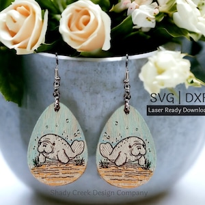 Puede incluir: Un par de pendientes de madera con un diseño de manatí nadando en agua turquesa cortado con láser. Los pendientes cuelgan de ganchos plateados y se muestran sobre una superficie gris con flores en el fondo. El texto "SVG | DXF Laser Ready Download" es visible en la imagen.