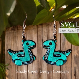 May include: A pair of turquoise blue dinosaur pool float earrings. The earrings are made of wood and have a shiny, glittery finish. The earrings are laser cut and ready to download. SVG | DXF Laser Ready Download. Shady Creek Design Company.