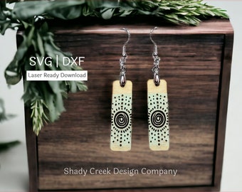May include: A pair of wooden earrings with a light green and natural wood finish. The earrings feature a black and white geometric design with a circle pattern. The earrings are hanging from silver hooks.