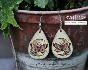 May include: A pair of wooden teardrop-shaped earrings with a laser-etched design of a moth and crescent moon. The earrings are painted white with a light brown wood grain showing through. The moth is a light pink and yellow colour with black markings. The crescent moon is a light yellow colour with a white star on it. The earrings are hanging from silver hooks.