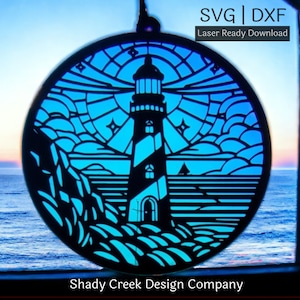 May include: A circular, laser-cut design featuring a lighthouse, rocks, and sea, viewed through a window. The design is black with a blue background, and the text "SVG | DXF Laser Ready Download" is visible. The words "Shady Creek Design Company" are also present.
