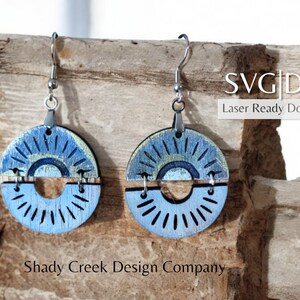 Puede incluir: Un par de pendientes de madera cortados con láser con un diseño de sol azul y verde. Los pendientes están hechos de dos piezas de madera que se unen con un anillo de metal. Los pendientes cuelgan de ganchos plateados. SVG|DXF Descarga lista para láser.