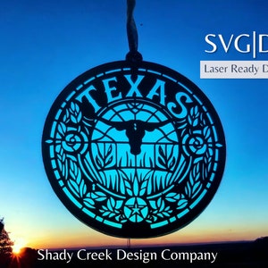 May include: A black and blue Texas ornament with a longhorn steer silhouette and floral designs. The ornament is hanging against a blue sky with a sunset in the background. The text "SVG|DXF Laser Ready Download" is in the upper right corner of the image. The text "Shady Creek Design Company" is at the bottom of the image.