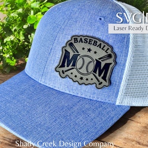 Puede incluir: Una gorra de béisbol azul y blanca con un diseño de mamá de béisbol en blanco y negro. El diseño presenta una pelota de béisbol, bates y estrellas. El texto "Baseball Mom" está escrito en un círculo alrededor del diseño.