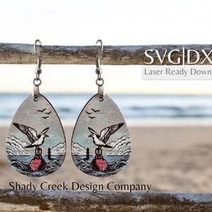 May include: A pair of wooden earrings with a seagull and buoy design. The earrings are teardrop shaped and have a silver hook. The background is a blurred image of a beach.