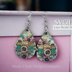 May include: A pair of teardrop-shaped wooden earrings with a vintage camera design and floral accents. The earrings are painted in shades of green, yellow, pink, and purple. The text "SVG|DXF Laser Ready Download" is visible on a pink surface behind the earrings.