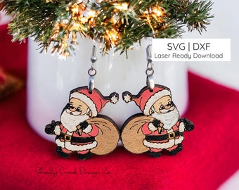 Op de afbeelding: Een paar houten oorbellen met een kerstman-ontwerp, rode muts, witte baard en bruine zak. De oorbellen zijn lasergesneden en klaar om te downloaden als SVG- en DXF-bestanden.