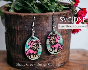 May include: A pair of wooden earrings with a bulldog wearing a pink bunny costume. The earrings are oval shaped and have a silver hook. The background is a wooden surface. SVG | DXF Laser Ready Download.