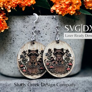 May include: A pair of wooden earrings featuring a laser-cut design of a raccoon sitting on a tree stump surrounded by mushrooms and flowers. The earrings are attached to silver hooks. The text "SVG|DXF Laser Ready Download" is visible in the image.