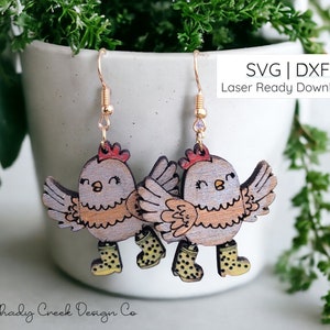 Puede incluir: Un par de pendientes de madera con un diseño de pollo de dibujos animados. Los pollos llevan botas de lluvia amarillas y tienen un esquema de color marrón claro y gris. Los pendientes están cortados con láser y listos para descargar como archivos SVG y DXF.