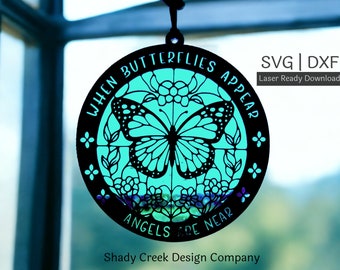 May include: A black and white circular design with the text "WHEN BUTTERFLIES APPEAR ANGELS ARE NEAR" and a butterfly in the center. The butterfly is outlined in black and filled with a teal color. The design is hanging from a string in front of a window.