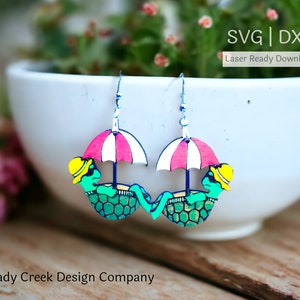 Może przedstawiać: Para laserowo wyciętych drewnianych kolczyków przedstawiających zielonego żółwia w żółtym kapeluszu, relaksującego się pod różowo-białym parasolem w paski. Kolczyki wiszą na białej misce. SVG | DXF Pobierz gotowy do cięcia laserem.