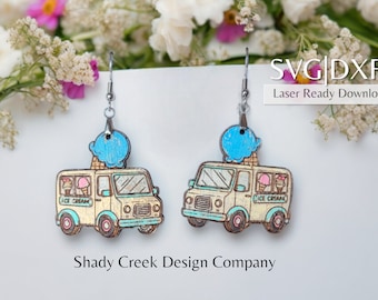 May include: A pair of wooden earrings featuring an ice cream truck with a blue ice cream cone on top. The truck is painted in a light yellow color with blue and pink accents. The earrings are attached to silver hooks.