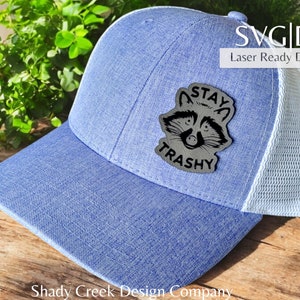 May include: A blue and white trucker hat with a black and white raccoon patch that says "Stay Trashy".