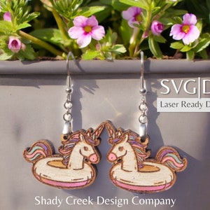 May include: A pair of wooden earrings featuring a unicorn riding an inflatable ring. The unicorn is white with a pink and purple mane and tail. The ring is white with a pink and purple stripe. The earrings are laser cut and ready to download as SVG and DXF files. Shady Creek Design Company.
