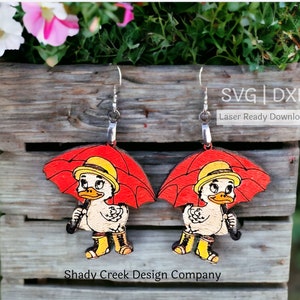 Op de afbeelding: Houten oorbellen met een cartoon eend met een gele hoed en laarzen, die een rode paraplu vasthoudt. De oorbellen zijn lasergesneden en hebben zilverkleurige haken. De tekst "Shady Creek Design Company" is zichtbaar.