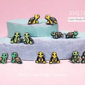 May include: SVG and DXF files for laser cutting turtle earrings. The image shows 8 pairs of turtle earrings in various colors, including green, yellow, and blue. The earrings are arranged on a light blue and pink background. The text "SVG | DXF Laser Ready Download" is visible in the top right corner of the image.