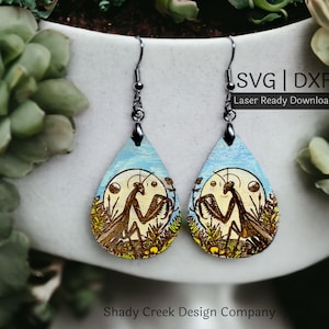 May include: A pair of wooden earrings with a blue background and a brown praying mantis design. The earrings are shaped like teardrops and have silver hooks. The text "SVG | DXF Laser Ready Download" is visible in the image.